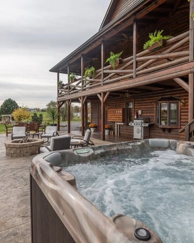 Hot tub on patio with log cabin, fire pit, lounge chairs, and pastoral landscape beyond
