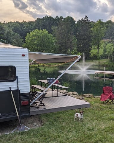 Travel trailer campsite with extended awning, camping chairs, fire pit, and lake view surrounded by forest.