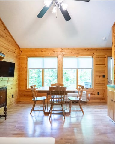Open-plan log cabin interior with kitchen, dining area, and large windows overlooking forest