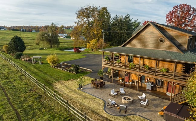 Log cabin with wraparound porch, patio with fire pit and lounge chairs, surrounded by rolling pastures and autumn trees.